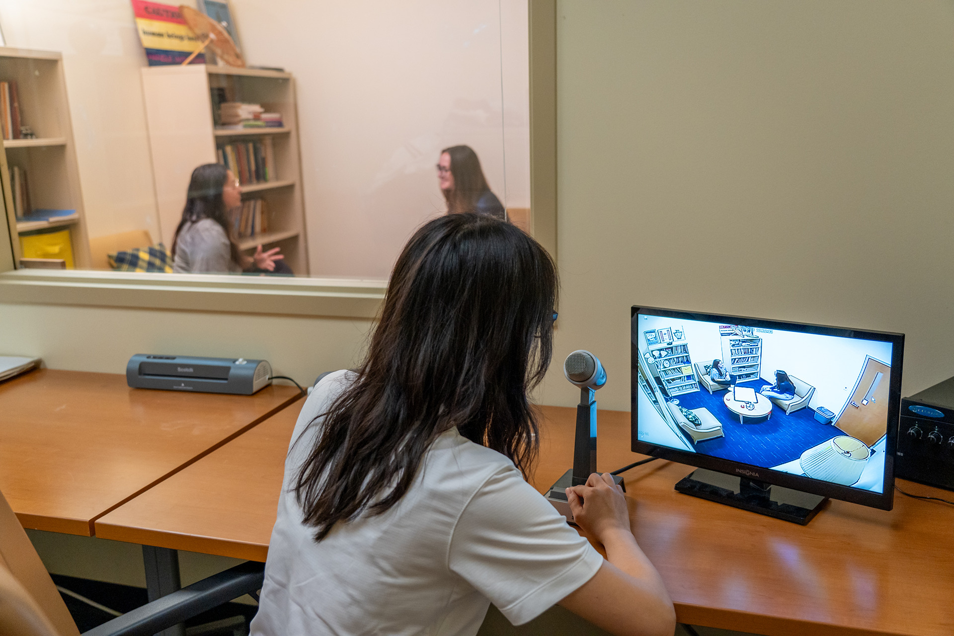 A student sits behind a two-way mirror observing a counseling session.