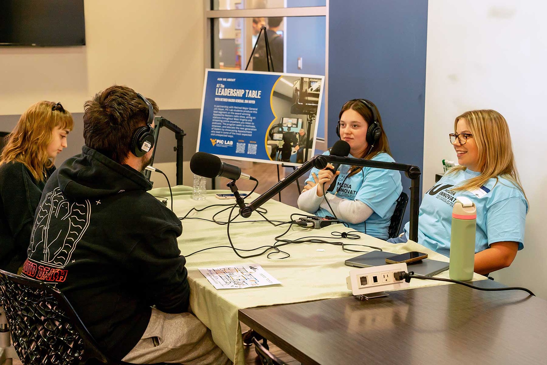 Four students seated around a table. Two are wearing headphones and speaking into microphones.