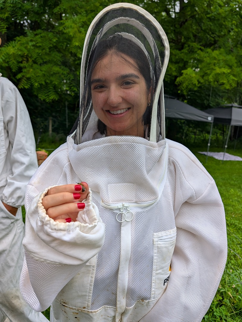 Amna Haque smiles while wearing a beekeeping suit outdoors.